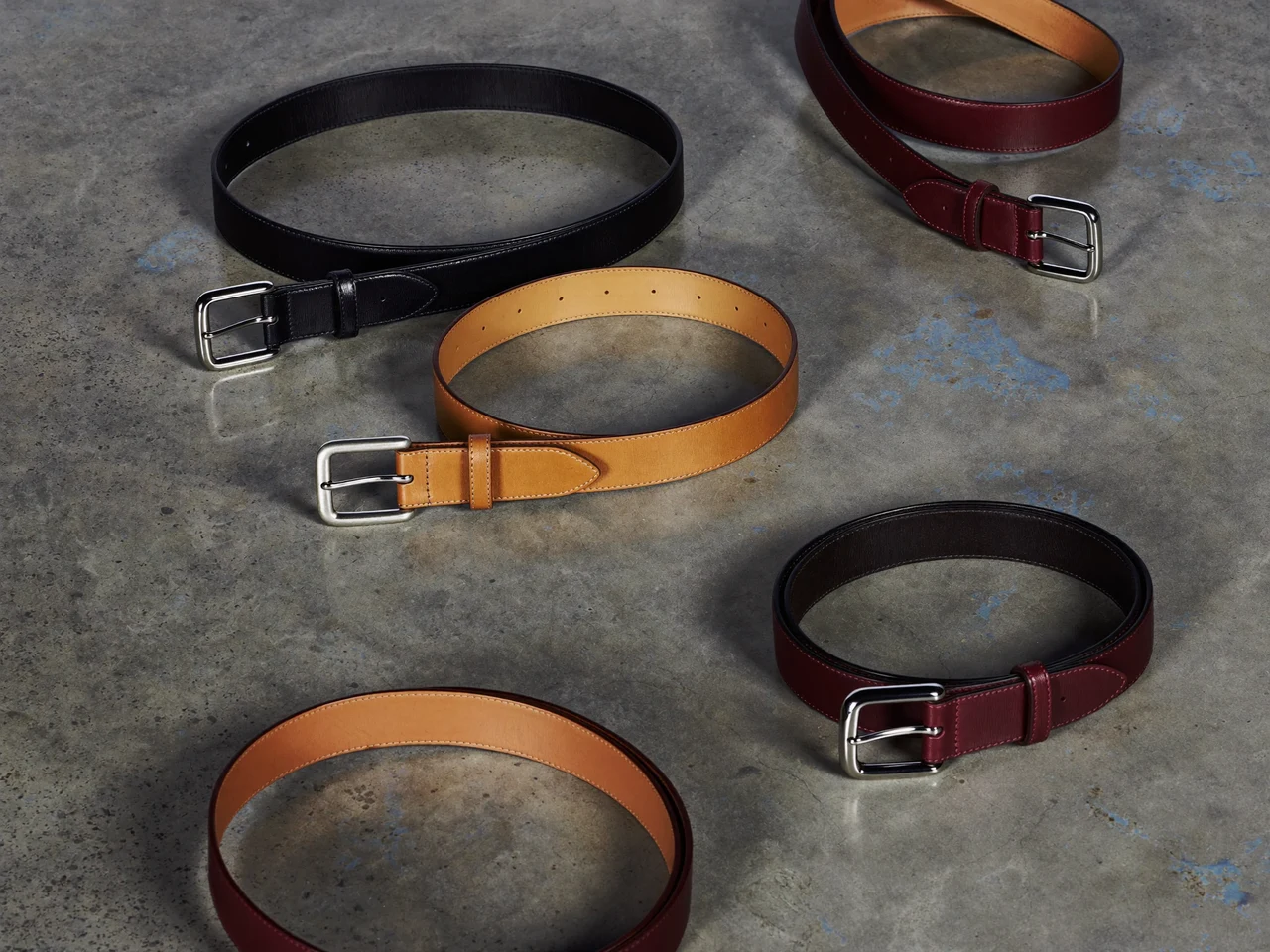 Curele negre, maro și bordo aranjate în evantai pe beton lustruit, cu reflexii reci.